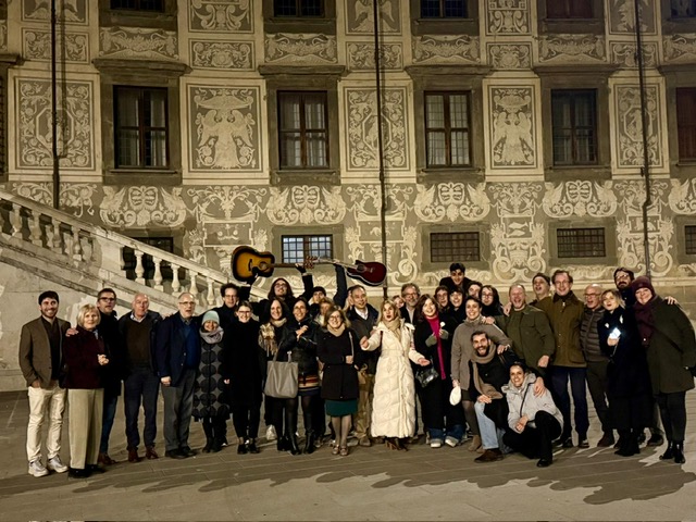 Participants of the MELAS Consensus standing in front of the Normale University in Pisa. There are about 40 people of mixed ages and genders wearing winter coats. It is evening and they are standing in front of a highly decorated Italian building. People at the back of the group are holding aloft two guitars. It looks as if people are cheering or singing in the photo.
