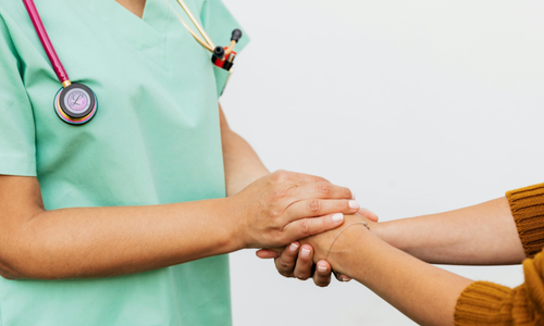 Photo of two people clasping hands. Only their hands, arms and one person's torso are visible. We can see the torso of the person on the left. They are wearing medical scrubs in light green, and have a stethoscope hanging over their neck. Their faces are not visible in the photo