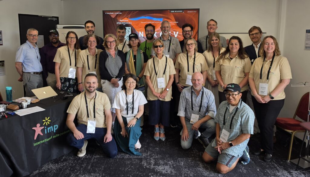Photo of a group of about 27 people crouching or sitting for a group photo. Most are wearing light brown t-shirts and lanyards. We can see a table with a IMP branded tablecloth to the left of the picture. 